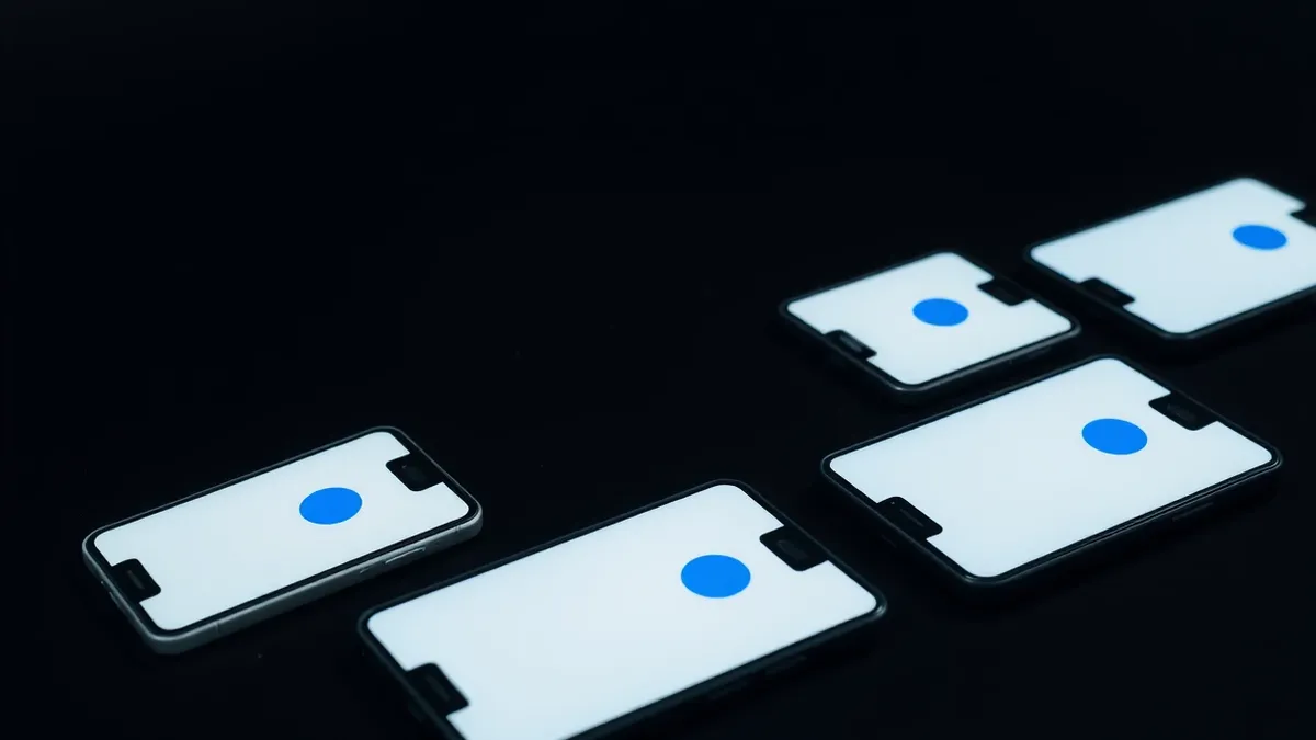 Illustration: A row of identical smartphones rests on a dark surface, each displaying a blank white screen with a single blue circular button.